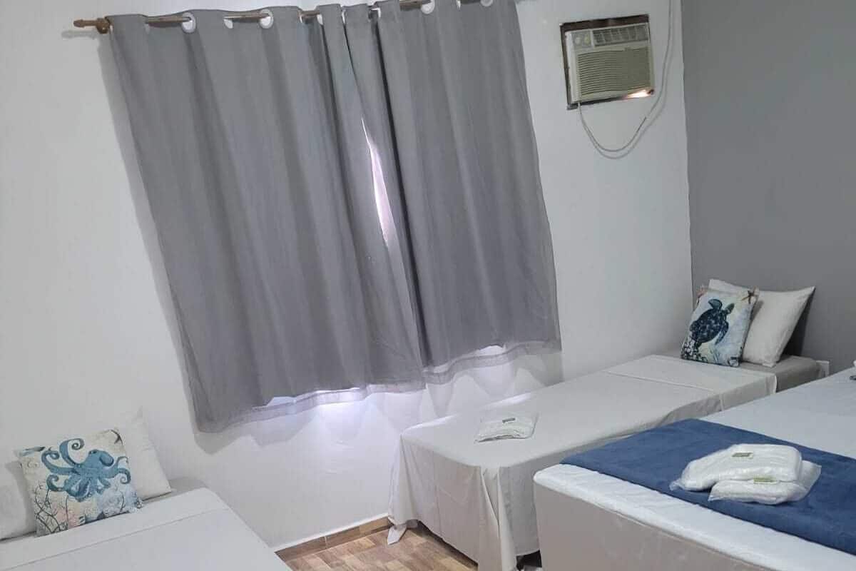 A hotel room with two single beds, a small table, a wall-mounted heater, and a window with grey curtains.