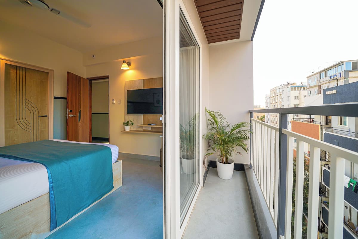 A balcony with a view of the city, a bed with a blue cover, a potted plant, and a small kitchen area.