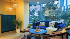 A modern living room with a blue sofa, two wooden chairs, a coffee table, and a large window offering a view of the cityscape. - Olive Hotel Kundalahalli by Embassy Group (Bengaluru)