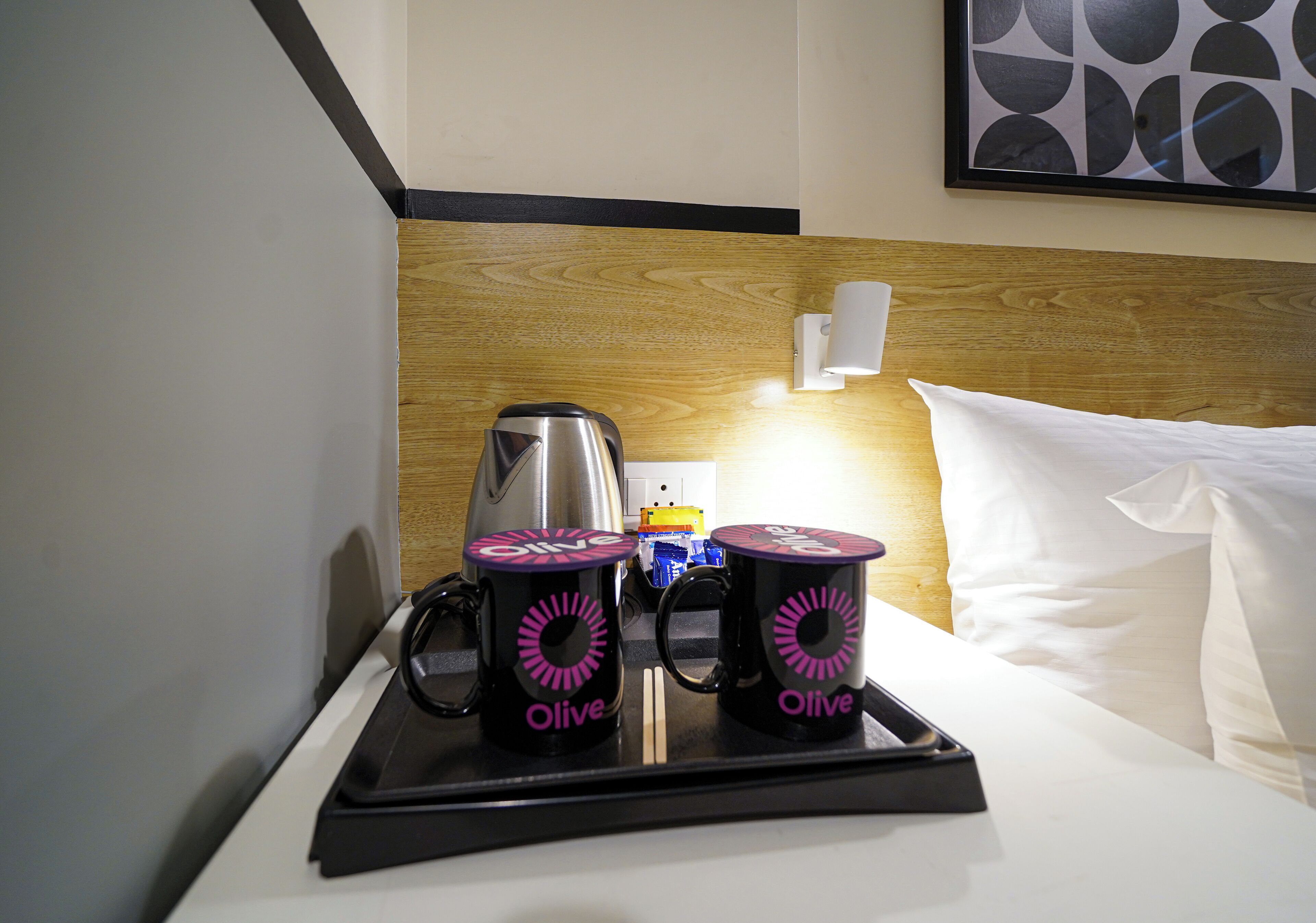 A hotel room with a wooden headboard, a bed with white pillows, a bedside table with a coffee maker and two mugs labeled "Olive."