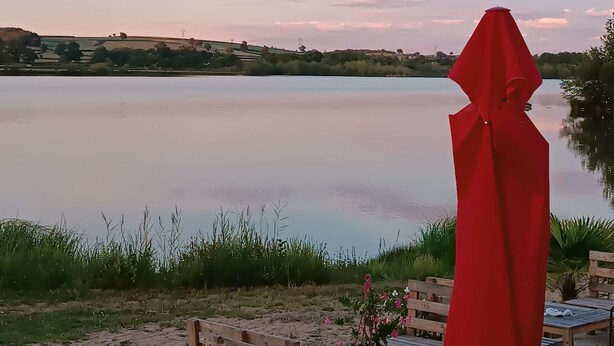 Un espace aménagé au bord du lac, avec des bancs et des tables en bois, un parasol rouge et une vue sur le lac et le paysage environnant.