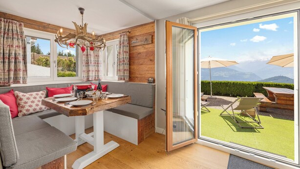 A cozy room with a wooden dining table set for two, a bench seating area, and a large sliding glass door leading to an outdoor patio with a view of mountains. - Cabane de la Delege -crans Montana- Amazing Views (Lens)