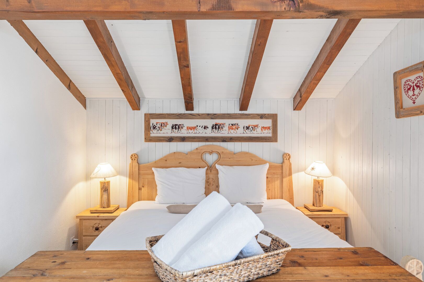 Une chambre confortable avec un lit en bois, du linge de lit blanc, deux lampes de chevet, un panier en osier contenant des serviettes et une œuvre d’art représentant des vaches, accrochée au mur.