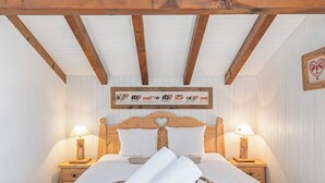 A cozy bedroom with a wooden bed, white linens, two bedside lamps, a woven basket with towels, and a wall-mounted artwork of cows. - Cabane de la Delege -crans Montana- Amazing Views (Lens)