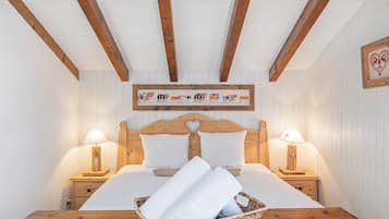 Une chambre confortable avec un lit en bois, du linge de lit blanc, deux lampes de chevet, un panier en osier contenant des serviettes et une œuvre d’art représentant des vaches, accrochée au mur.