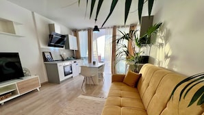 A modern living room with a sofa, a TV, a kitchen area, and a dining table. - Apartamento Julia (Costa Adeje)