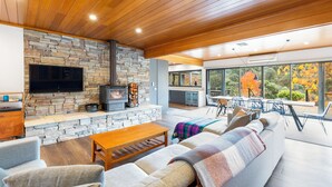 A modern living room with a stone fireplace, a flat-screen TV, and a large window overlooking a garden. - Delatite Valley: 4  BR, 3.5  BA House in Merrijig, Sleeps 12 (Merrijig)