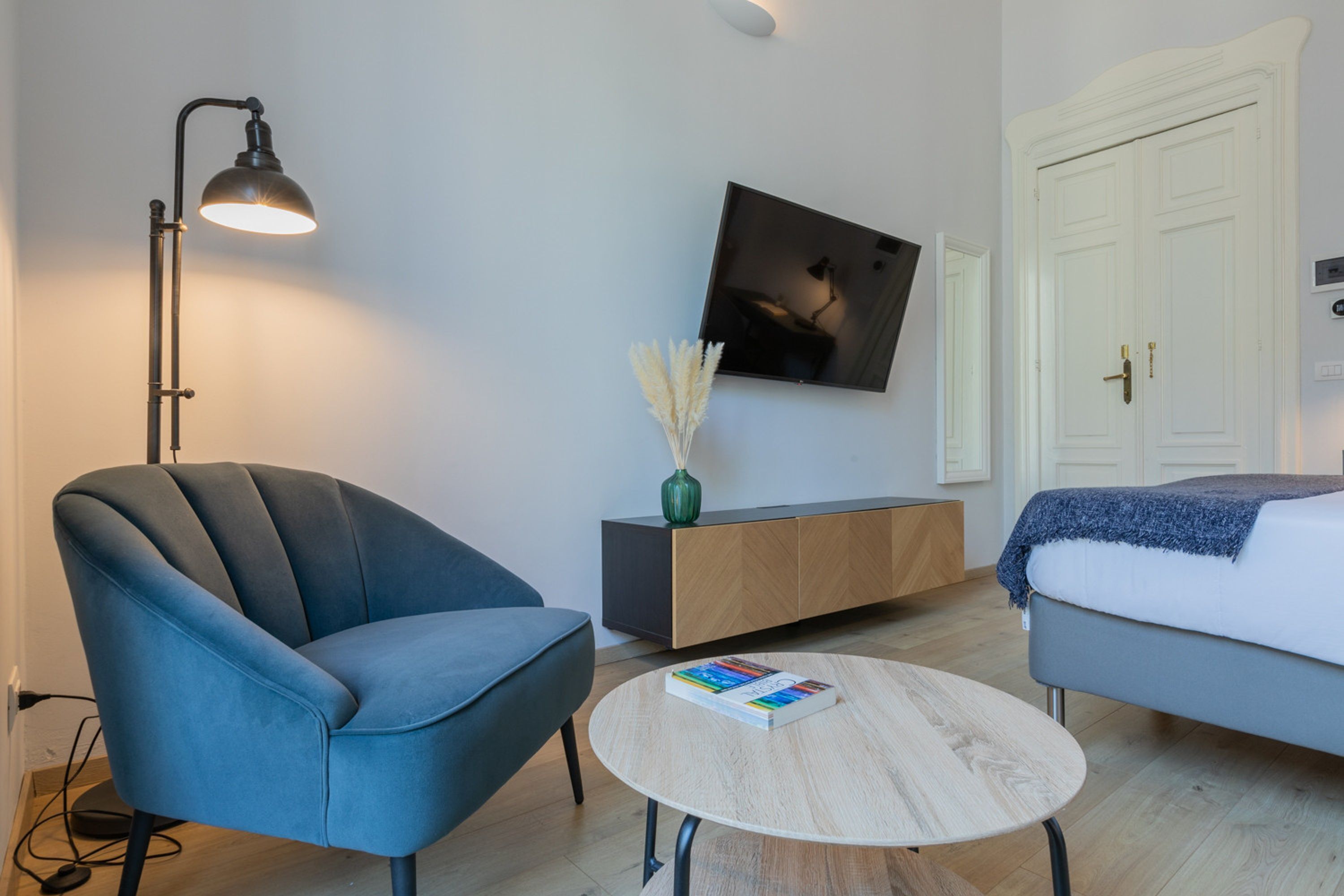 Une pièce moderne avec un fauteuil bleu, une table basse ronde sur laquelle repose un livre, un lampadaire, un téléviseur fixé au mur et une armoire blanche.
