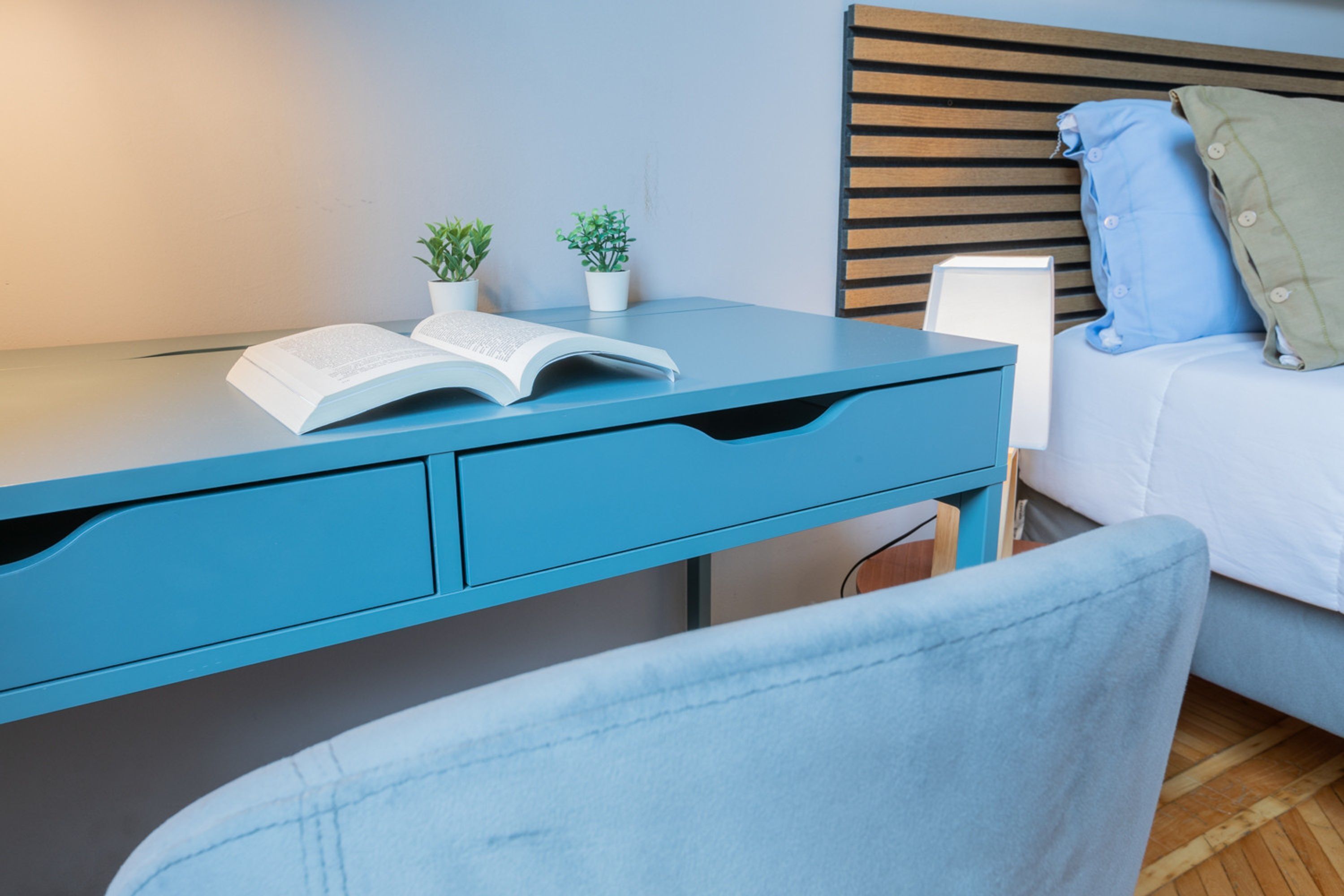 Um quarto moderno com uma escrivaninha azul, uma cama com roupa de cama branca e uma pequena planta em vaso na escrivaninha.