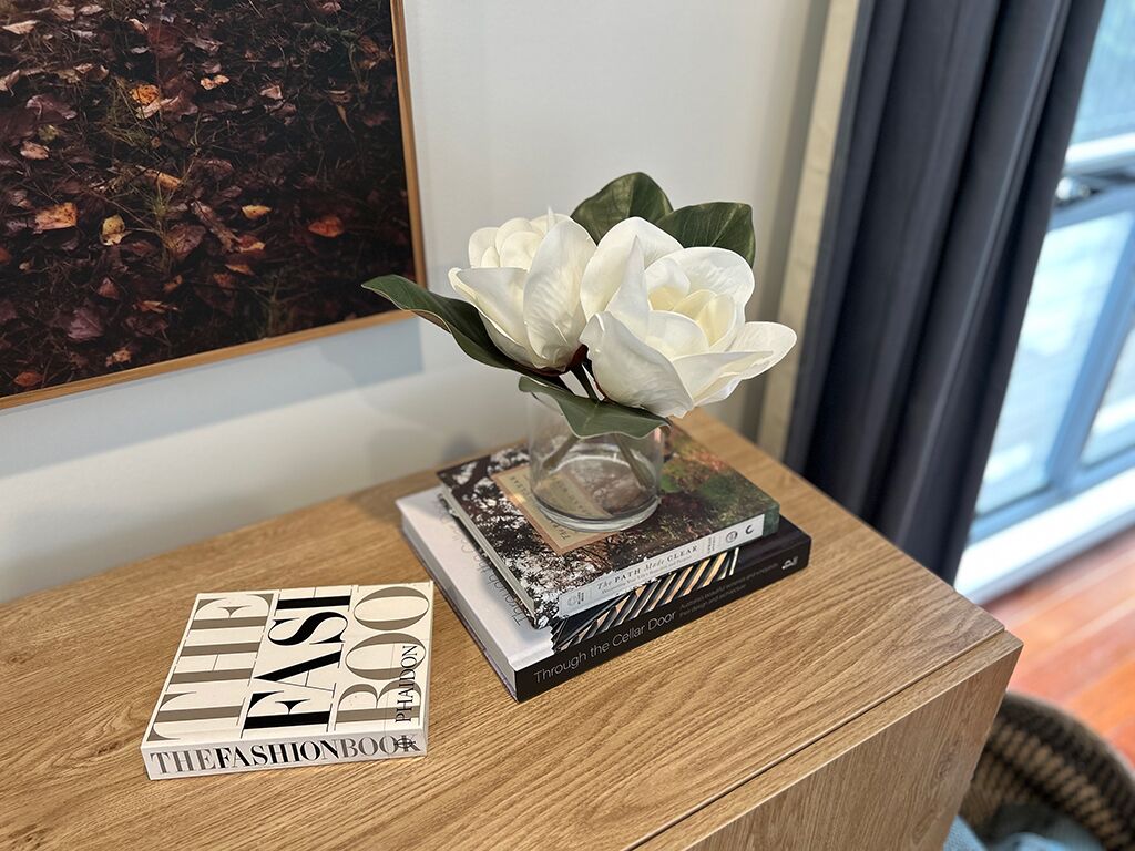 Une petite table d’appoint en bois, sur laquelle se trouve un vase de fleurs blanches, un livre intitulé « Le livre de la mode » et un autre livre intitulé « À travers la porte du col ».