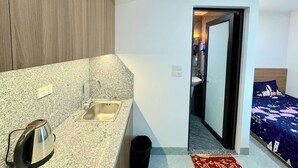 A compact hotel room with a kitchenette, a bed with a patterned bedspread, and a small bathroom visible through an open door. - Ruby Sun Home (Hanoi)