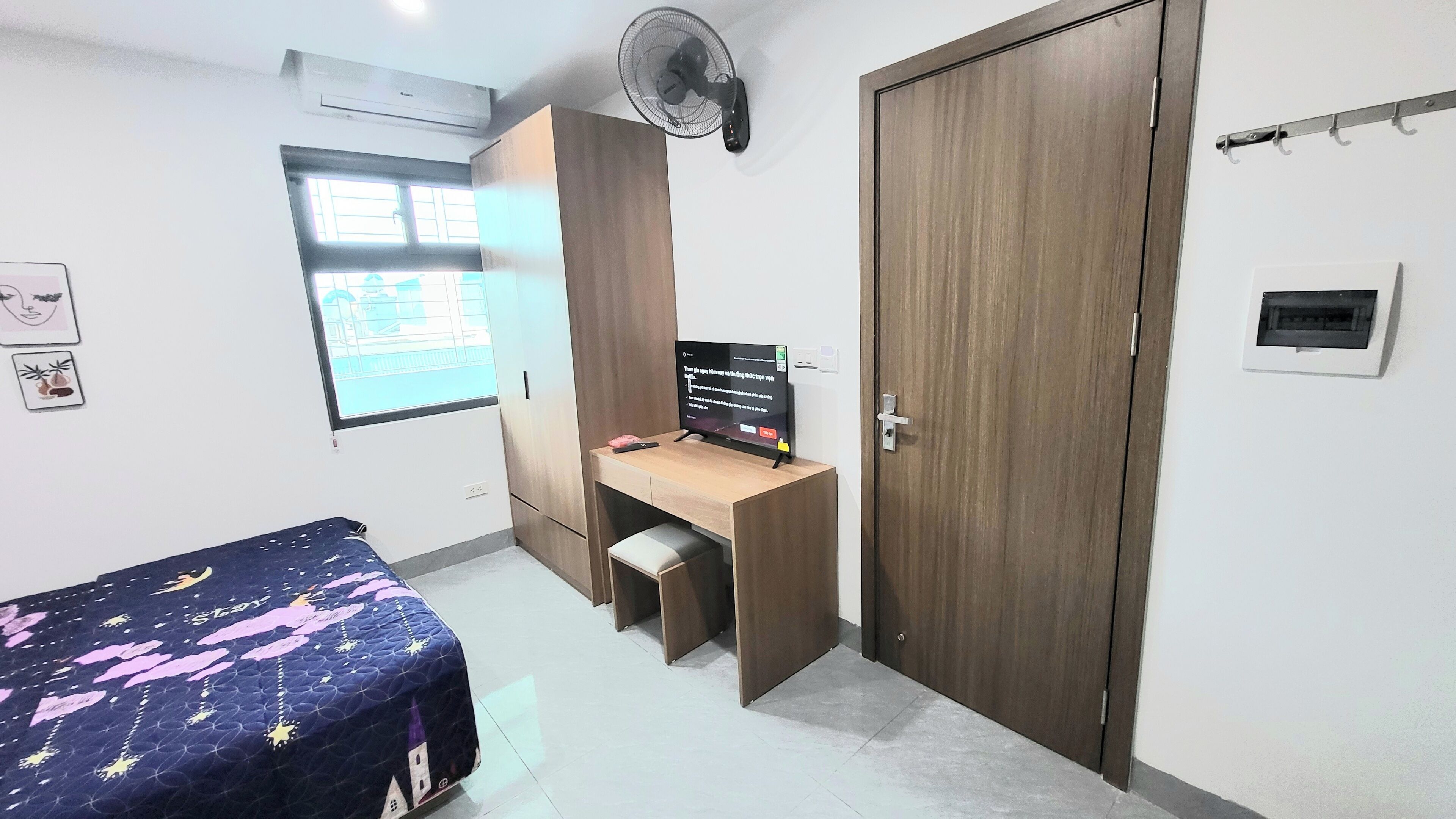 A bedroom with a bed, a desk, a wardrobe, a fan, and a window.
