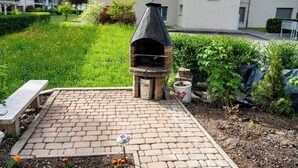 Une terrasse en briques avec un four extérieur en pierre, entourée de verdure et agrémentée d’un banc.
