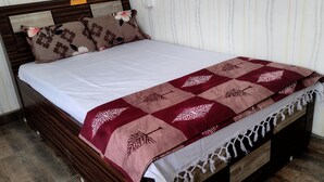 Una cama con sábana blanca y manta color burdeos con estampado de hojas, flanqueada por almohadas florales, en una habitación con suelo de madera y paredes blancas.