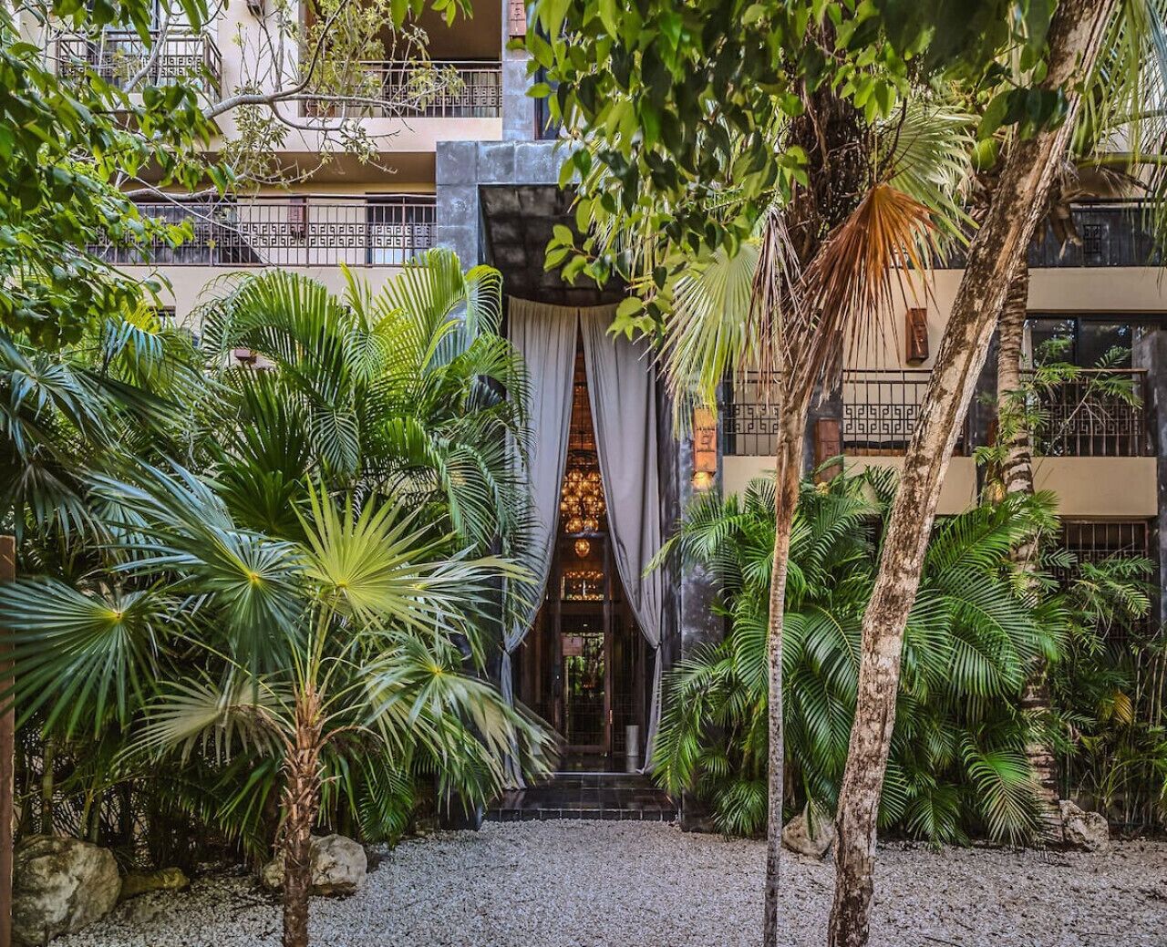 L’entrée d’un hôtel, avec un passage couvert, des palmiers et la façade d’un immeuble ornée de balcons.