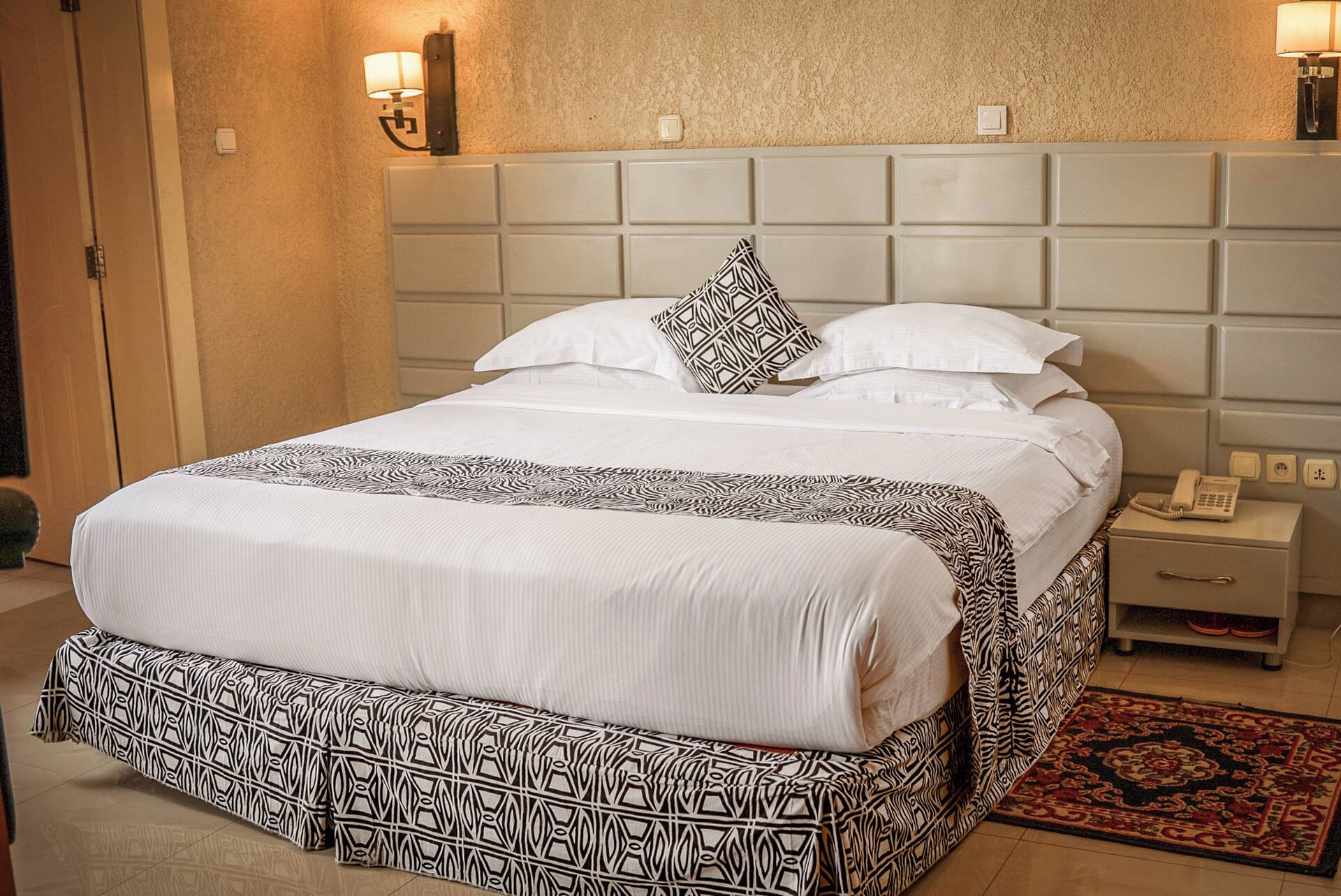 A hotel room with a large bed, a bedside table with a lamp, and a patterned rug.