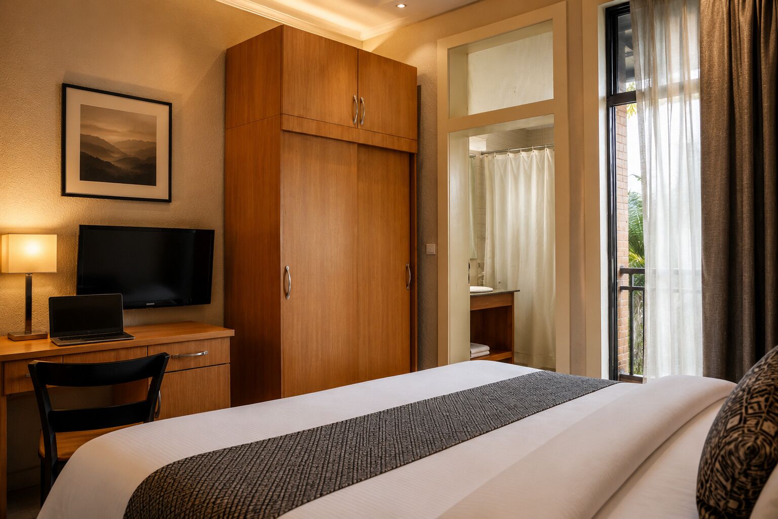A hotel room with a bed, a desk with a laptop, a television, and a large window with curtains.