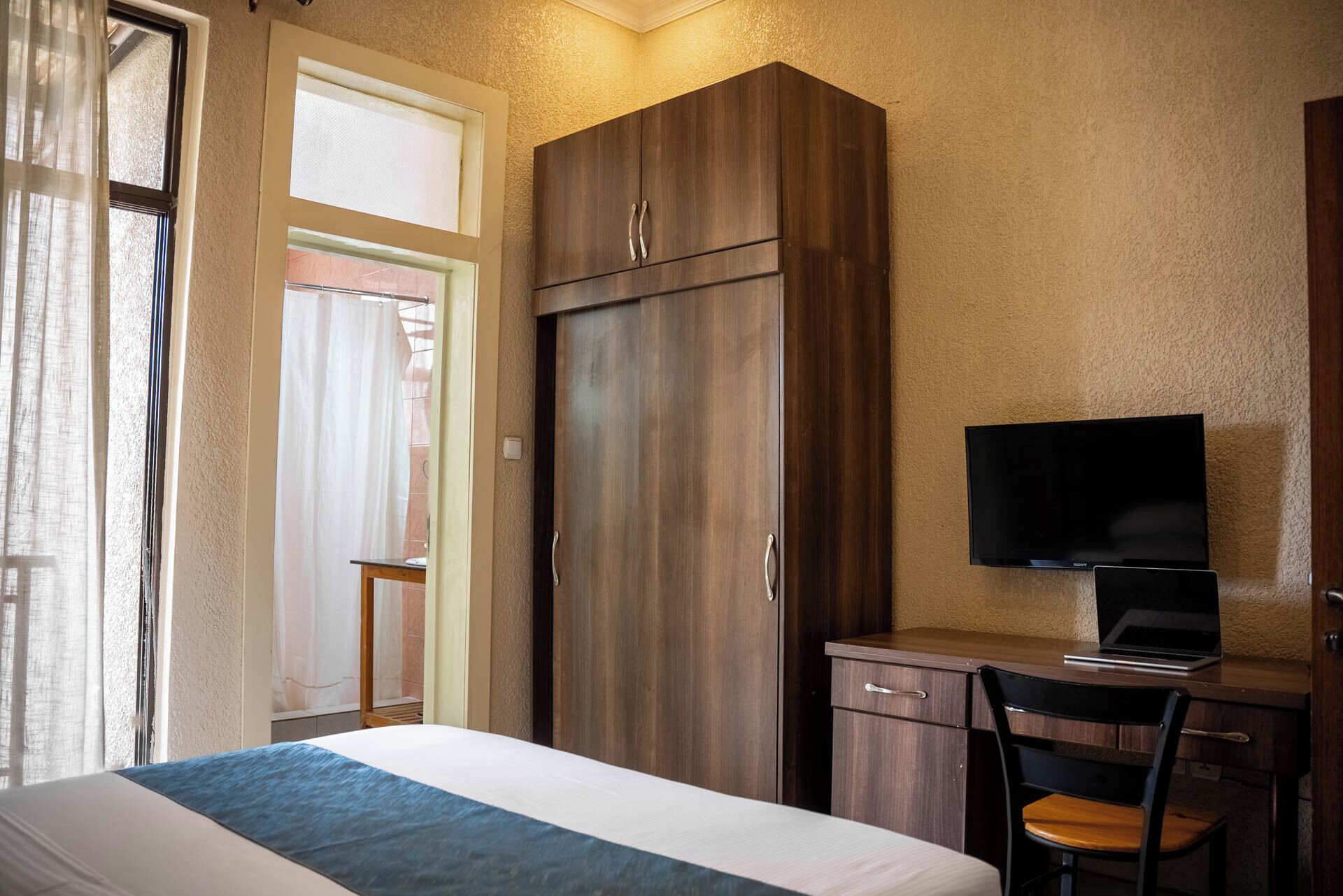 A hotel room with a bed, a desk with a chair, a wardrobe, a television, and a window with curtains.