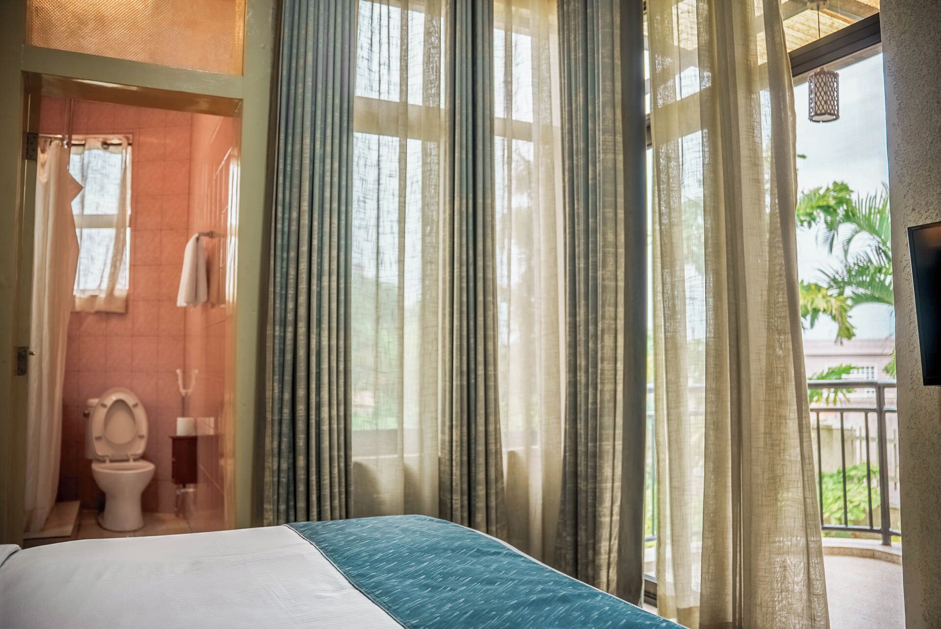 A hotel room with a bed, a toilet, and a balcony with curtains.