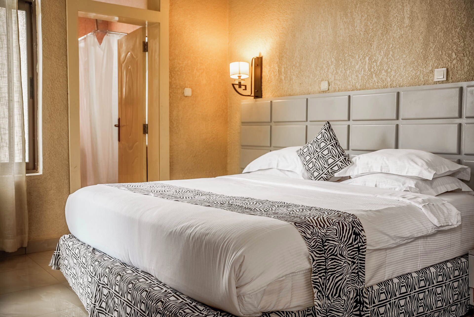 Ein Hotelzimmer mit einem großen Bett, einem Kopfteil, einer Wandleuchte und einer Tür mit Vorhang.