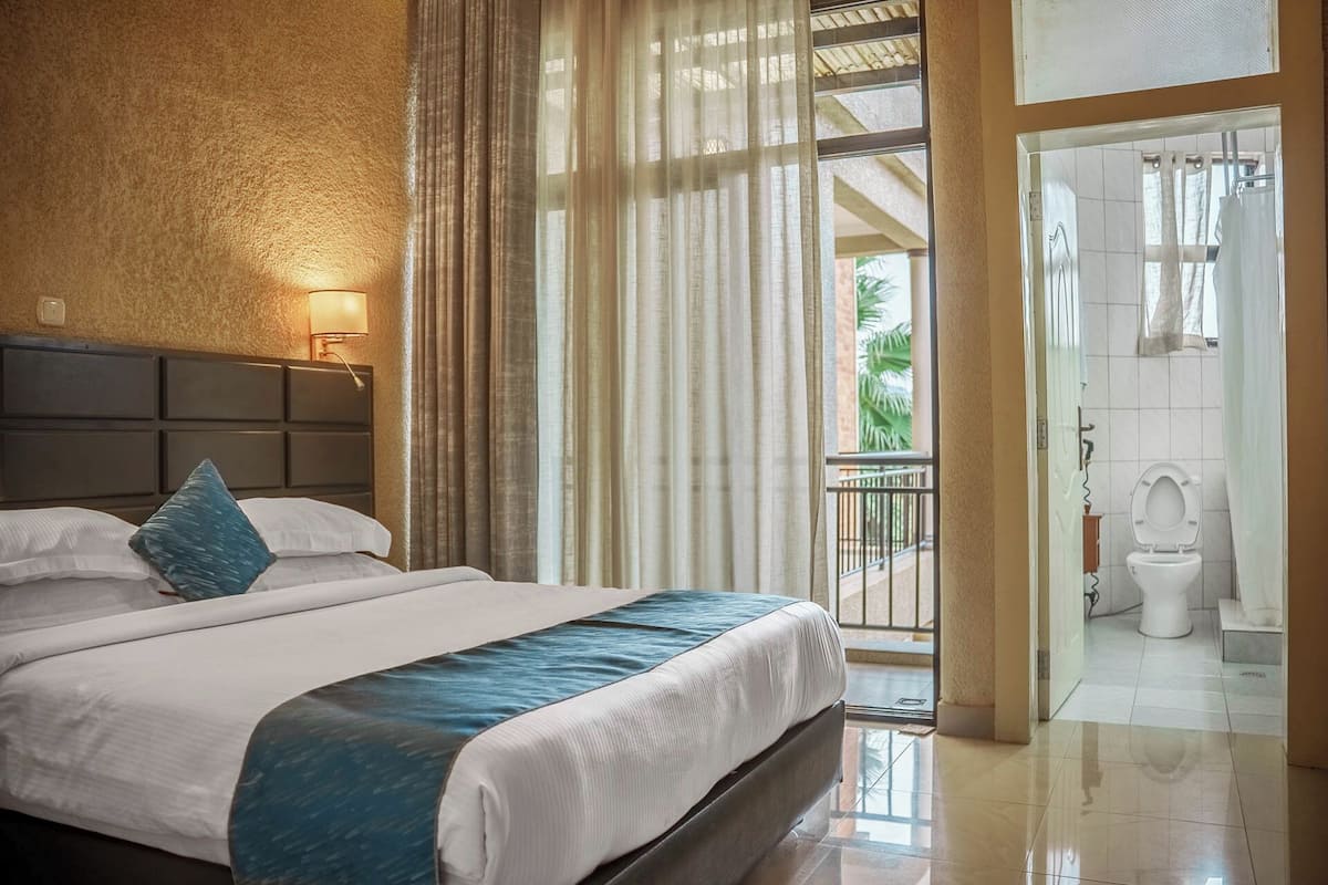 Habitación de hotel con una cama grande, una mesita de noche con lámpara, vista al balcón exterior y un baño con inodoro y lavabo.
