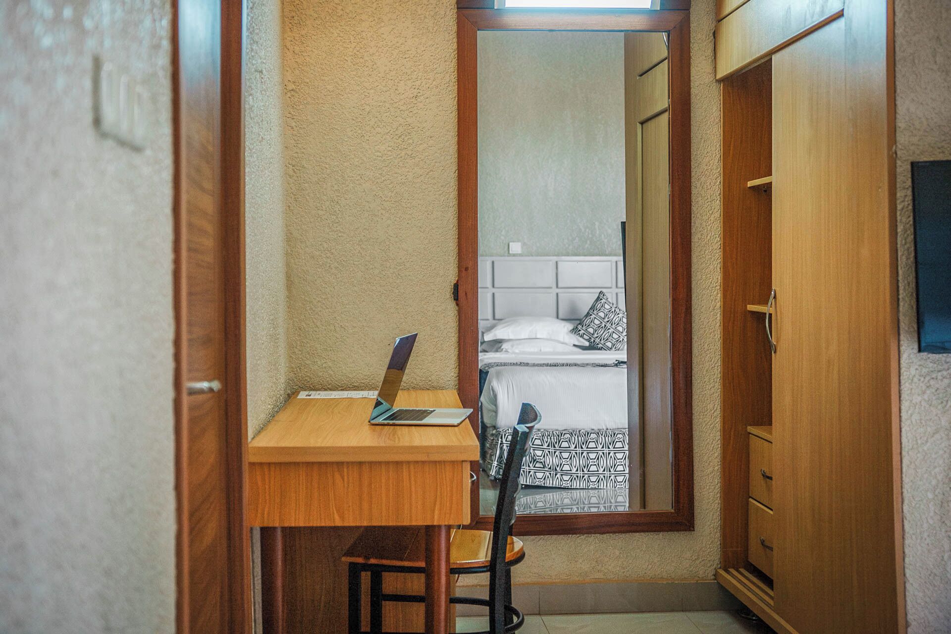 Una stanza piccola e stretta con una scrivania di legno, una sedia e un letto con copriletto decorato. Un portatile è sulla scrivania.