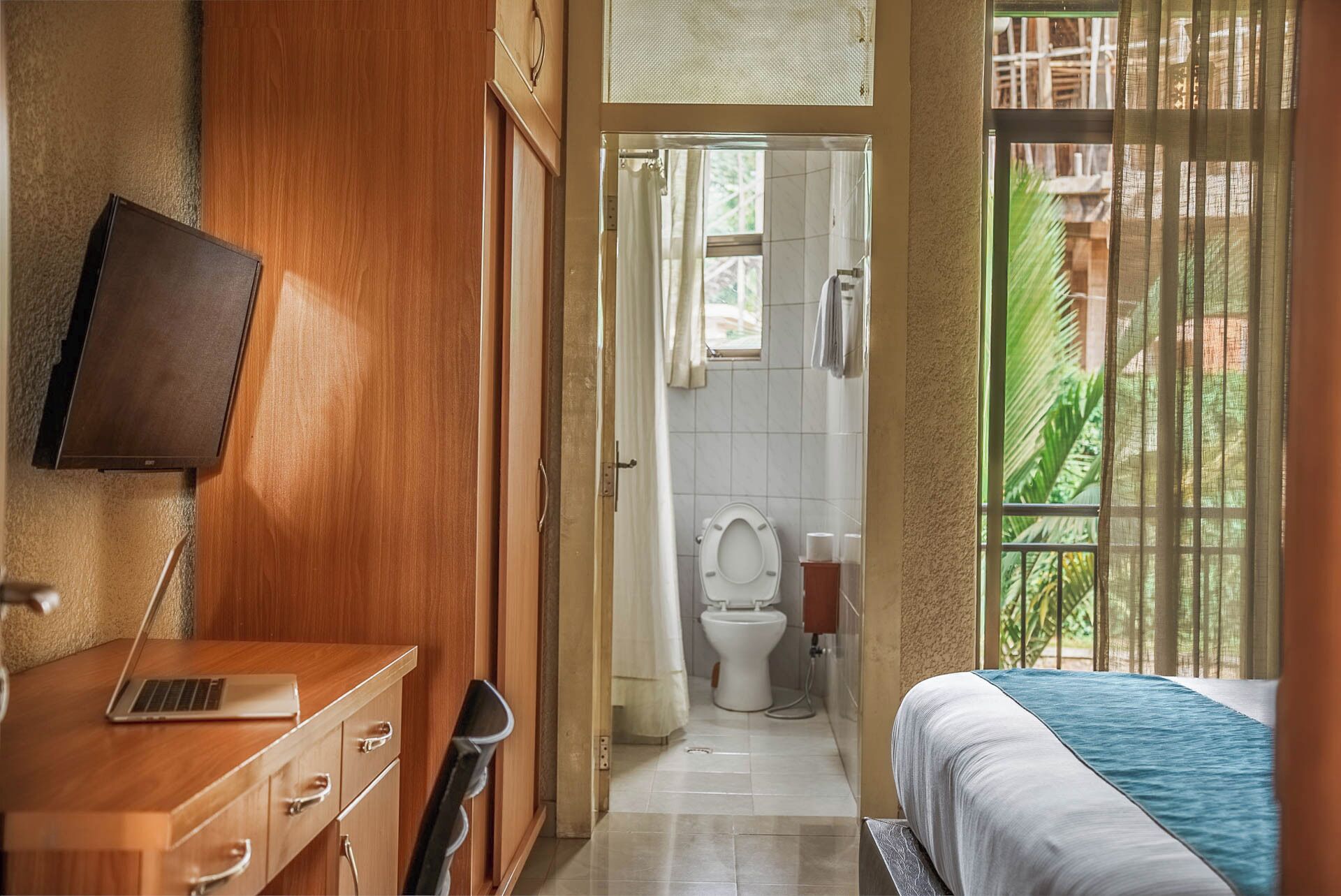 A bedroom with a bed, a wooden desk with a laptop, a toilet, and a shower area.