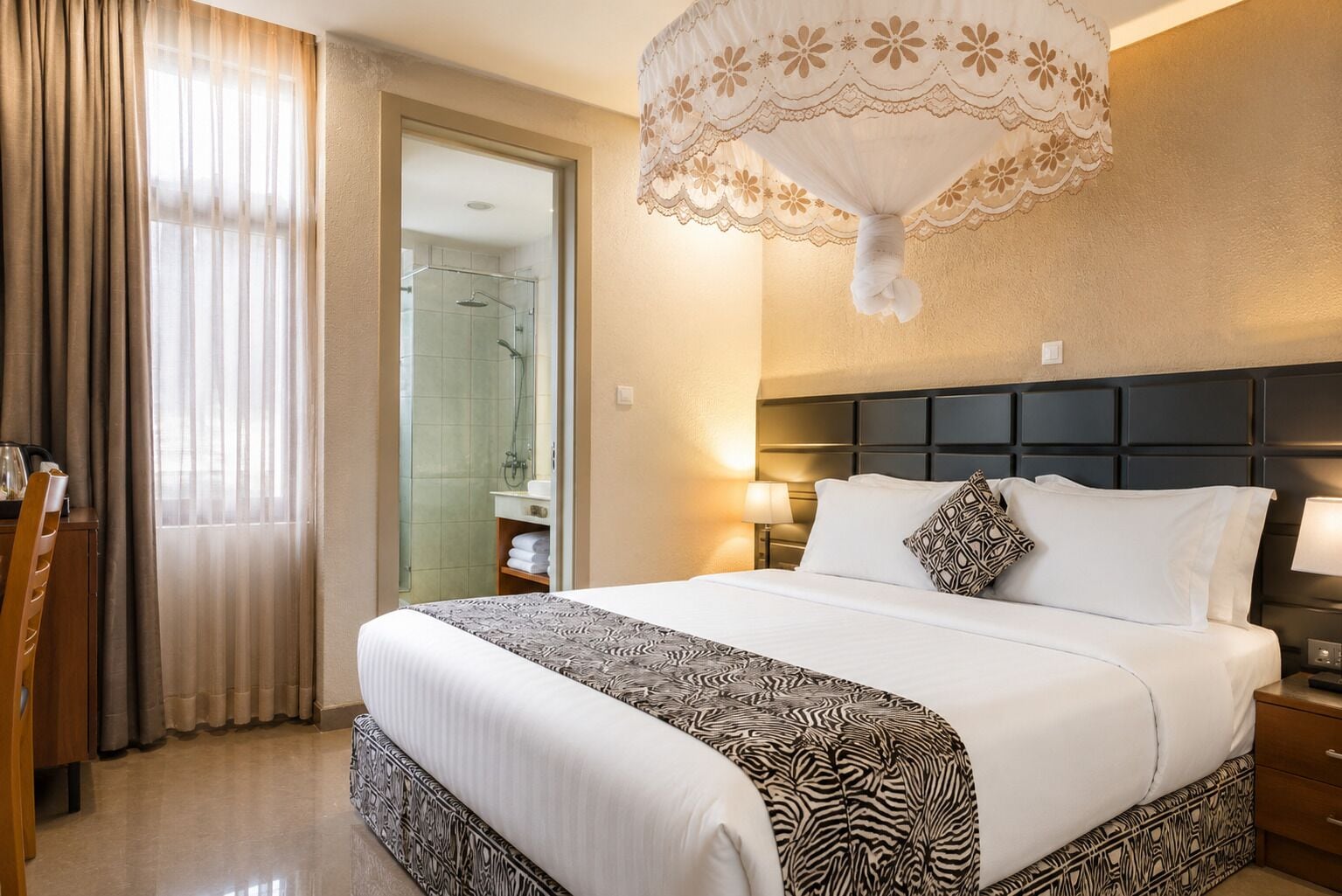A hotel room with a bed, bedside tables, a nightstand, a lamp, a shower, and a window with curtains.