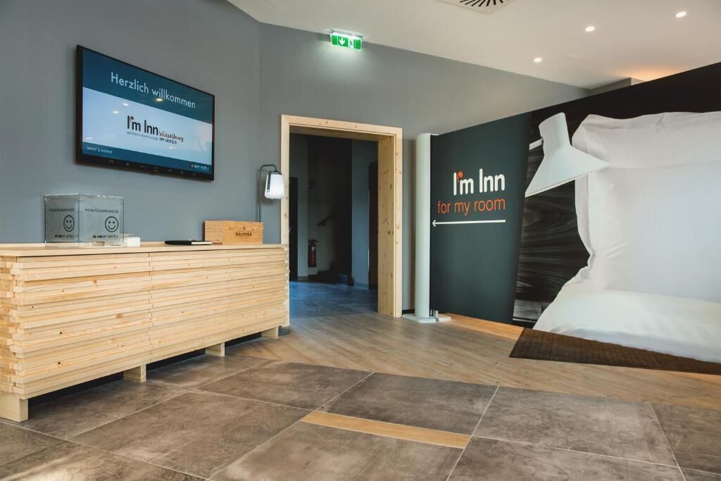 A hotel reception area with a wooden reception desk, a television displaying a welcome message, and a large bed with a pillow and a blanket.