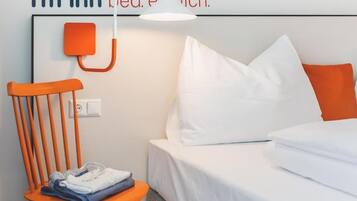 A hotel room with a bed, white bedding, an orange chair, and a wall-mounted lamp.