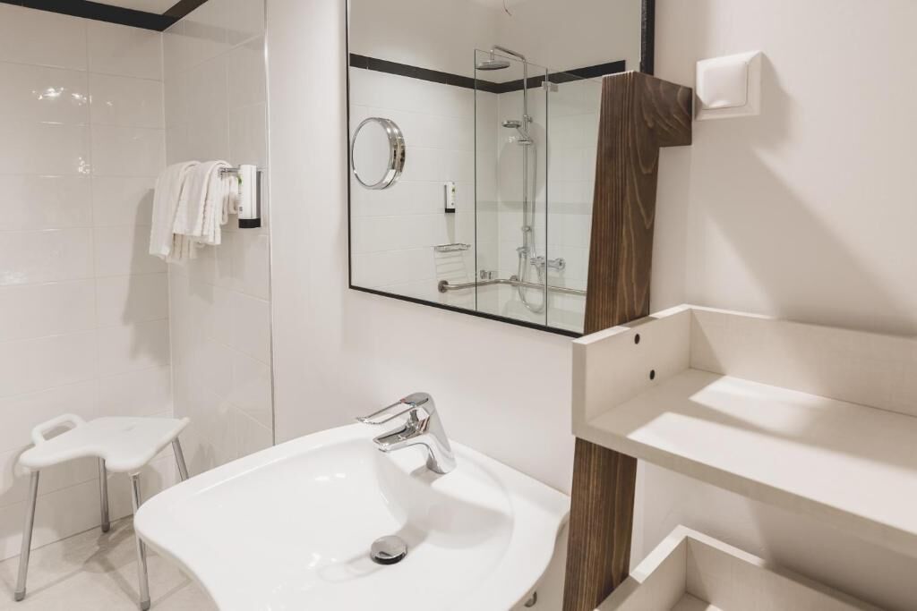 A modern bathroom with a large mirror, a glass-enclosed shower, a white sink, and a small stool.