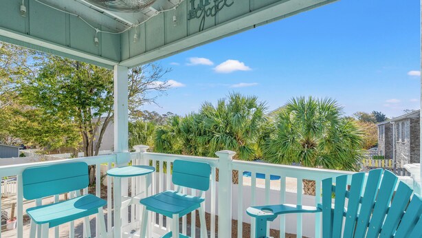 Un espace extérieur aménagé avec une zone de repos couverte, dotée de chaises Adirondack turquoise et d’une petite table, offrant une vue sur un jardin avec des palmiers et un ciel d’un bleu limpide.