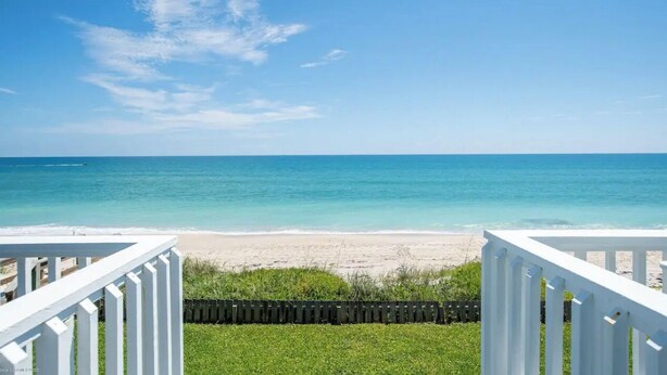 A beach view from a deck with white railings, clear blue sky, and calm sea.