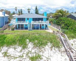 Une maison à deux étages avec un balcon, entourée d’une clôture en bois et donnant sur une plage de sable.