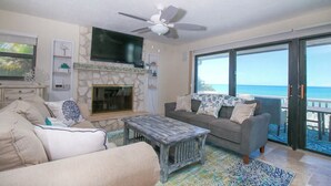 A living room with a fireplace, a ceiling fan, a large window with a view of the ocean, and a sofa with decorative pillows.