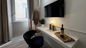 Ein modernes Wohnzimmer mit einem an der Wand befestigten Flachbildfernseher, einem Holztisch mit einer Weinflasche und einem Glas, einem dunkelblauen Sessel, einer kleinen Topfpflanze und einem Fenster mit Vorhängen.