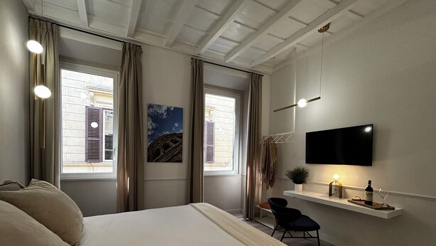 Ein Schlafzimmer mit einem Bett, einem an der Wand montierten Fernseher, einem kleinen Schreibtisch mit Stuhl und einem Fenster mit Vorhängen.