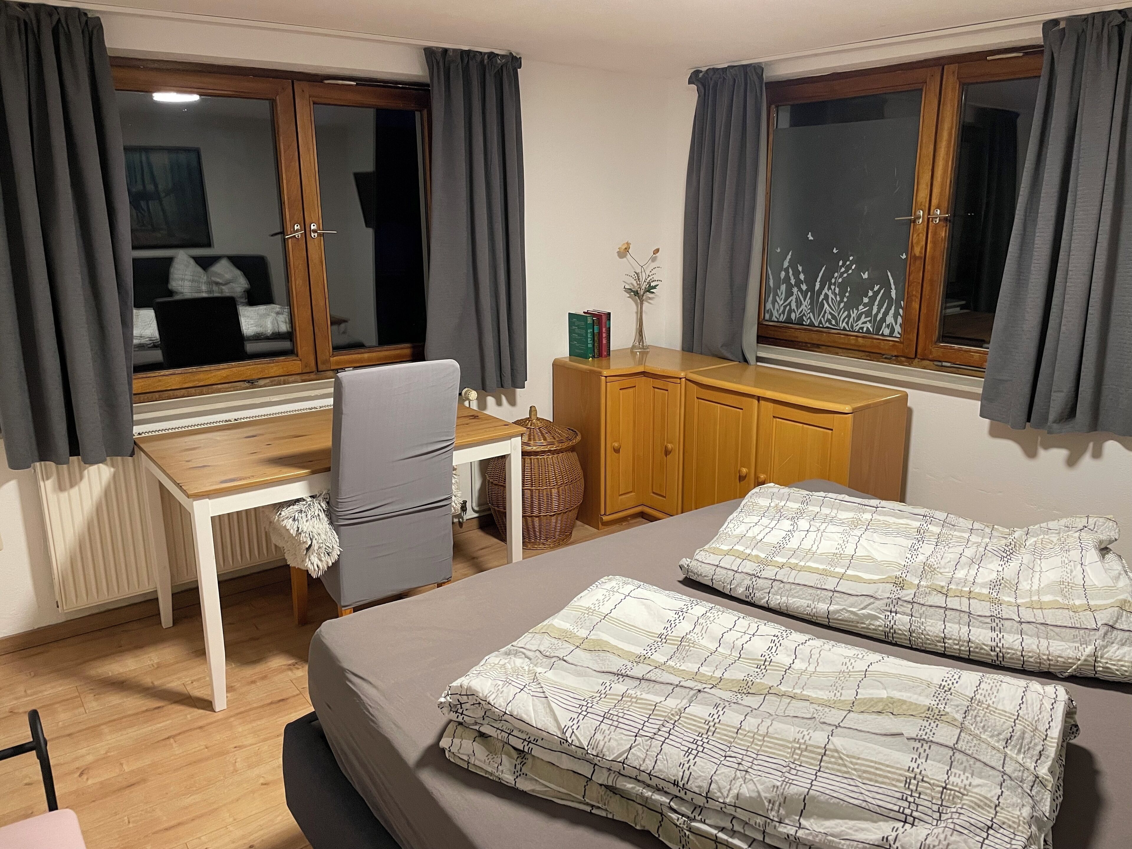 Une chambre d’hôtel avec un lit, un bureau et une chaise. Il y a une fenêtre avec des rideaux, un radiateur et une petite table sur laquelle est posé un vase.