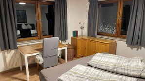 A hotel room with a bed, a desk, and a chair. There is a window with curtains, a radiator, and a small table with a vase. - Holiday Home with Private Bar and Sauna (Duderstadt)