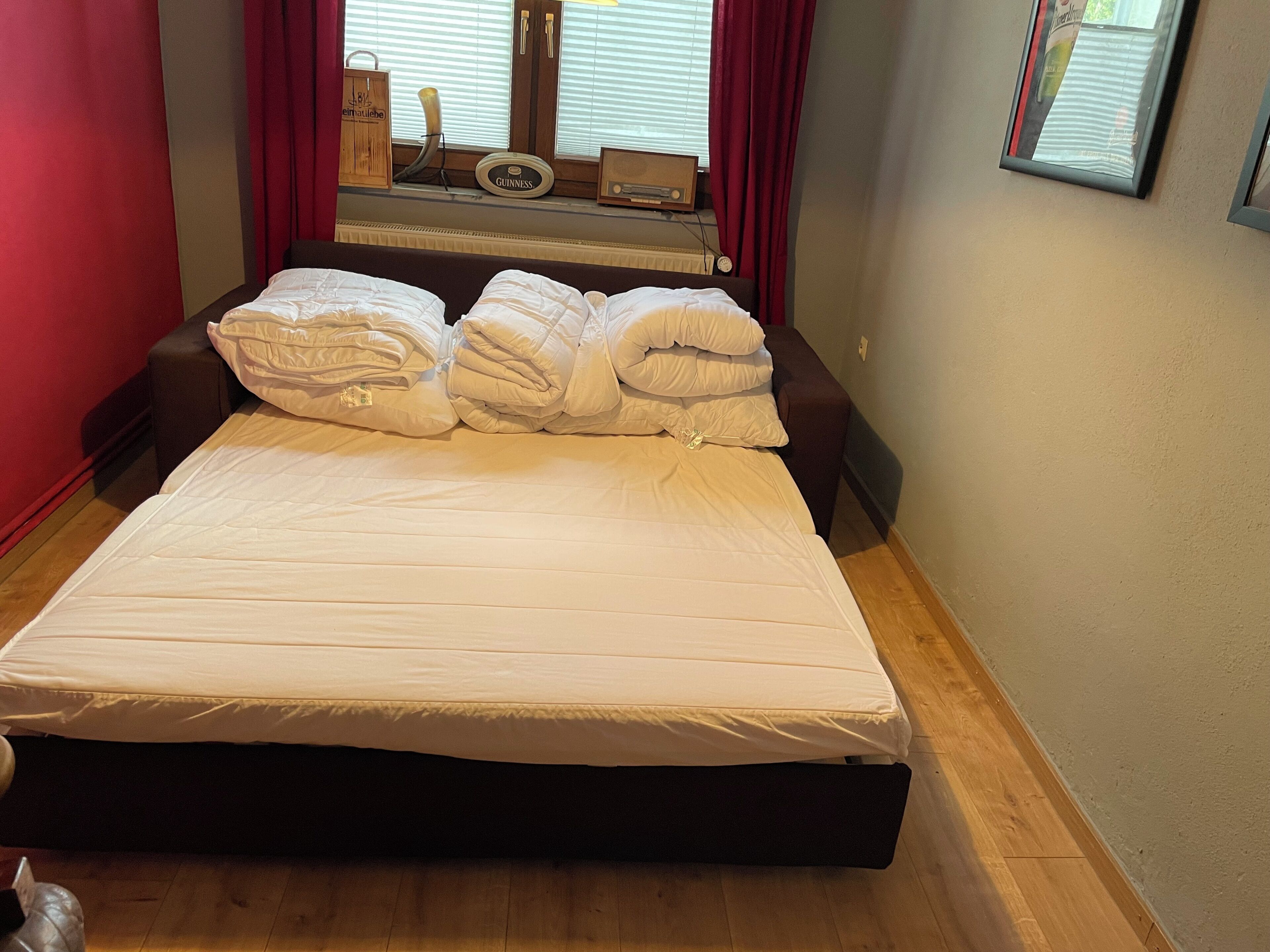 A neatly made bed with white bedding and multiple pillows, a wooden headboard, and a window with blinds and curtains.