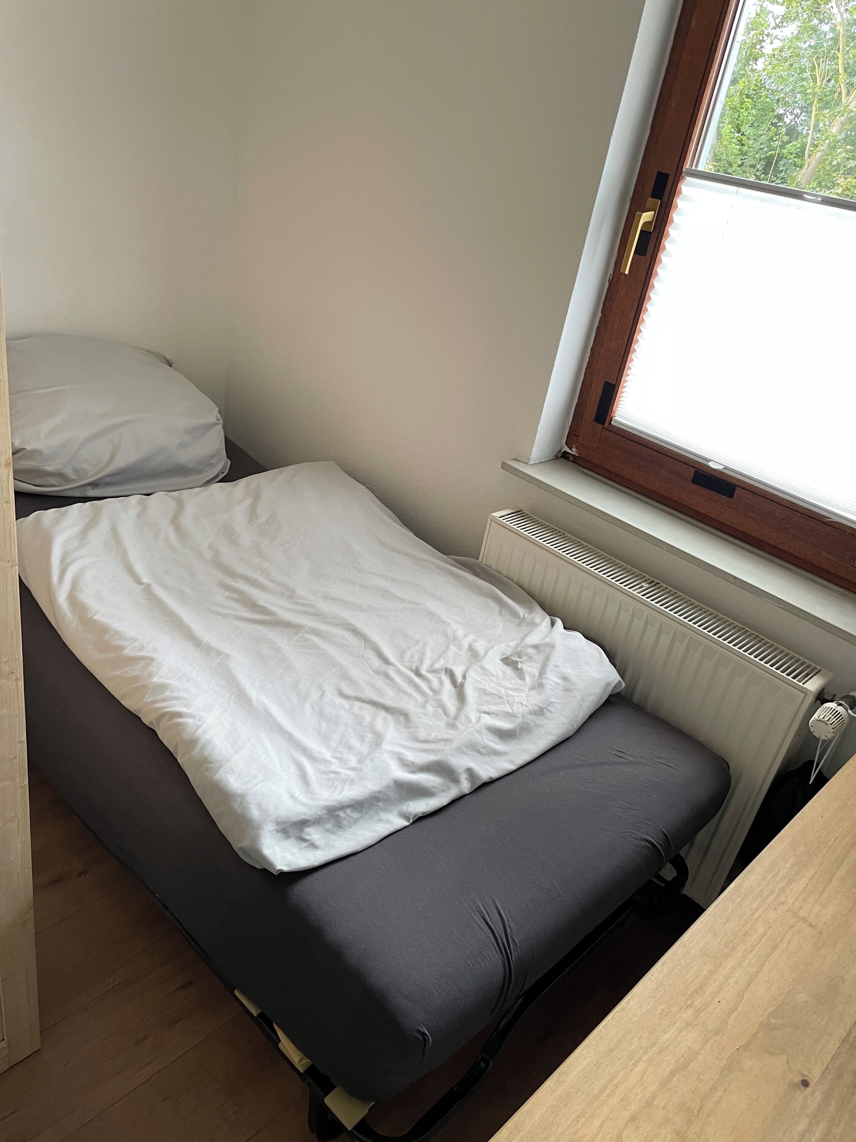A small, single-bed room with a window, a radiator, and a wooden table.