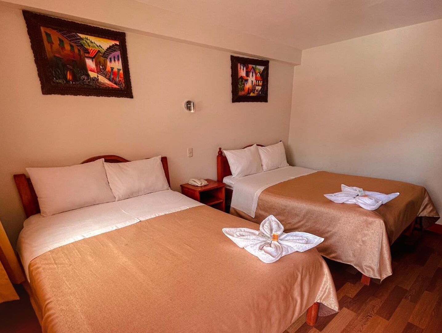 Habitación de hotel con dos camas individuales, cada una con una colcha marrón y almohadas blancas. Dos cuadros decoran la pared sobre las camas, y una mesita de noche con un teléfono encima.