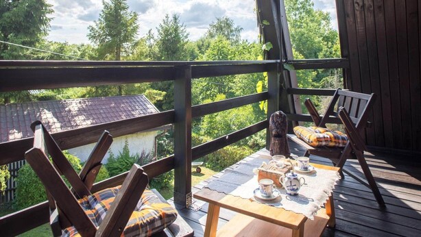 A wooden deck with a table set for tea, surrounded by chairs with plaid cushions, overlooking a forested area.