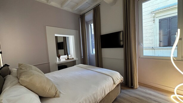 A hotel room with a bed, a bedside table with a mirror, a flat-screen TV mounted on the wall, and a window with curtains. - Suites Pantheon Rome - 3 (Roma)