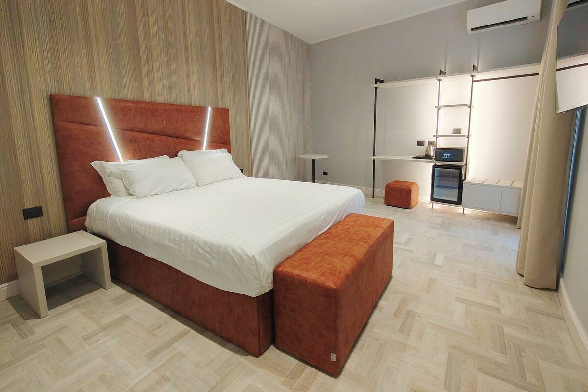Moderni hotellihuone, jossa on suuri sänky, yöpöytä, pieni jakkara, työpöytä tuolilla ja ilmastointilaite.