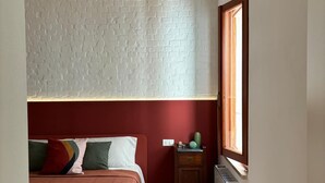 Une chambre avec un mur décoré d’un motif en briques, un lit avec une tête de lit rouge, une table de chevet ornée d’un cactus et une fenêtre offrant une vue agréable.