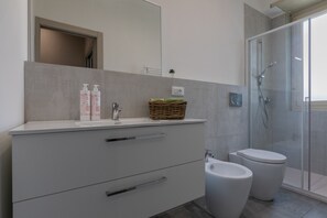 Ein modernes Badezimmer mit einem weißen Waschtisch, zwei weißen Toiletten, einer Duschkabine aus Glas und einem kleinen Blumentopf auf dem Waschtisch.