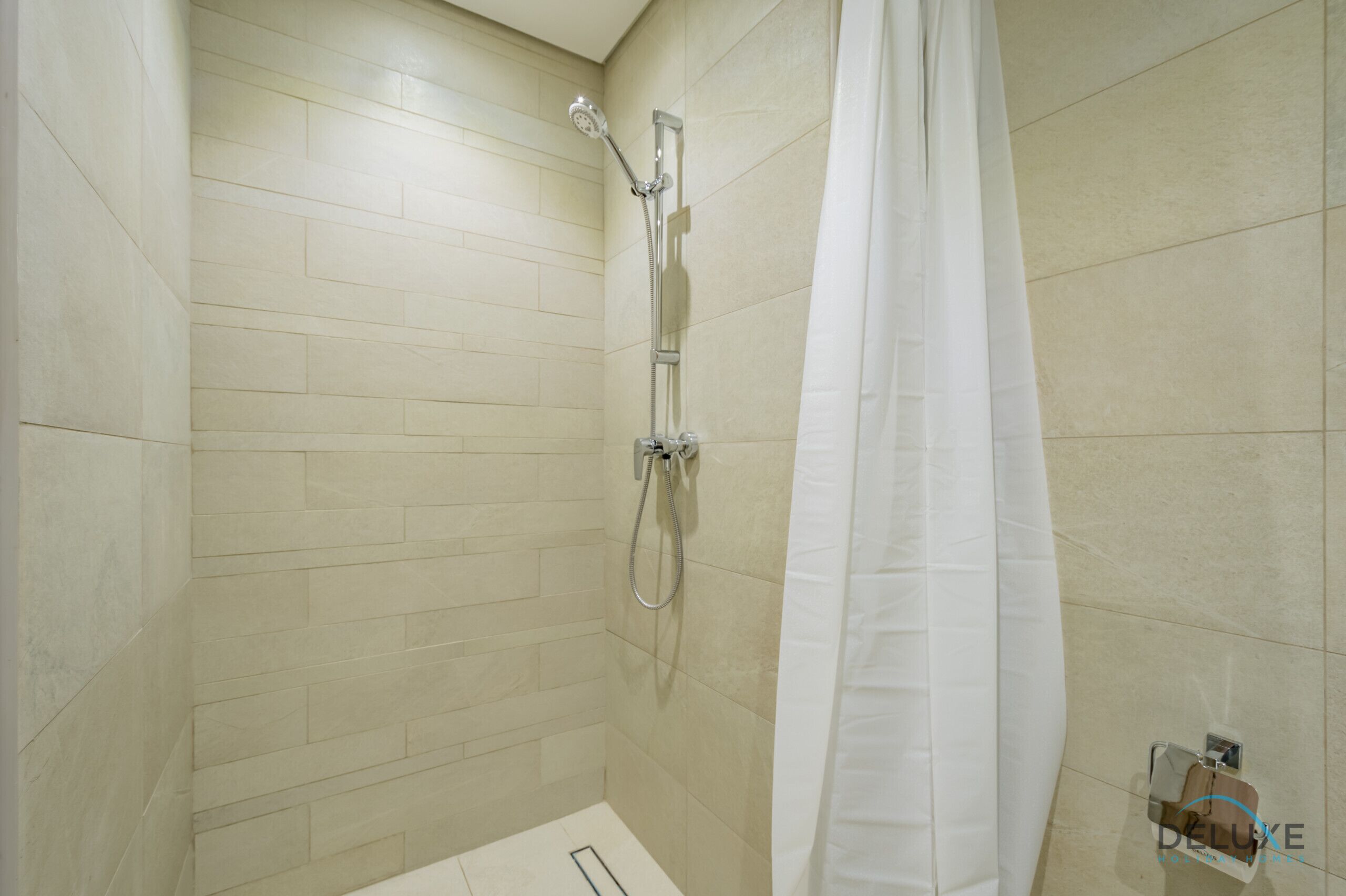 Een badkamer met beige betegelde muren, een douche met een handdouche en een wit douchegordijn.