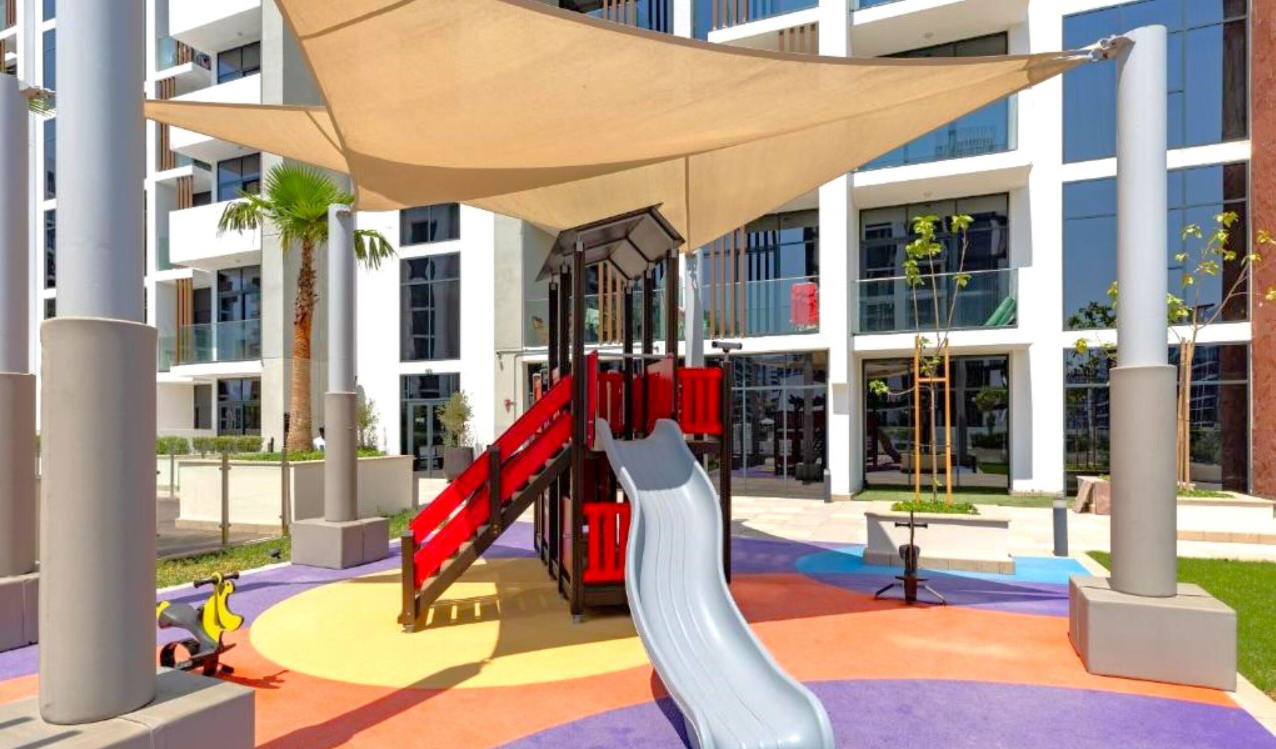 A playground with a slide and climbing structures, surrounded by modern buildings with large windows.