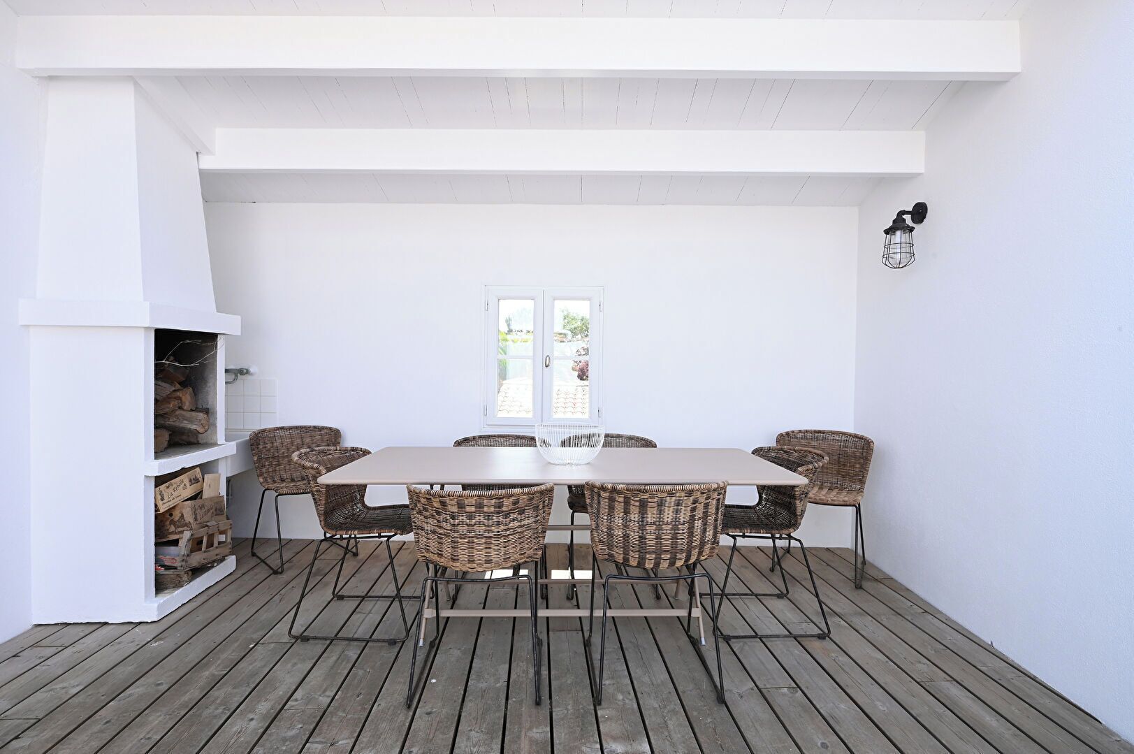 A covered patio with a wooden table and wicker chairs, a white fireplace, and a view through a glass door.